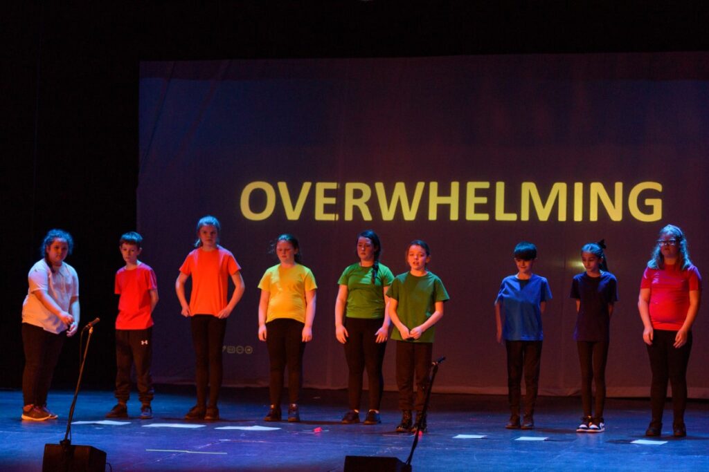 A group of young people wearing different coloured t shirts standing in a line on stage in front of a screen displaying the word 'overwhelming.'