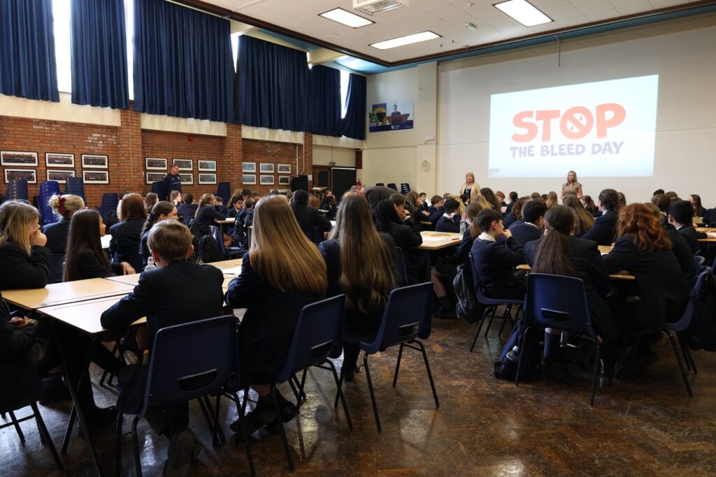 Pupils from Salesian Academy of St john Bosco at a session in school for Stop the bleed day. They are all sitting around tables looking towards a teacher at the front of the room. A large screen is displaying the wording Stop the bleed day. 